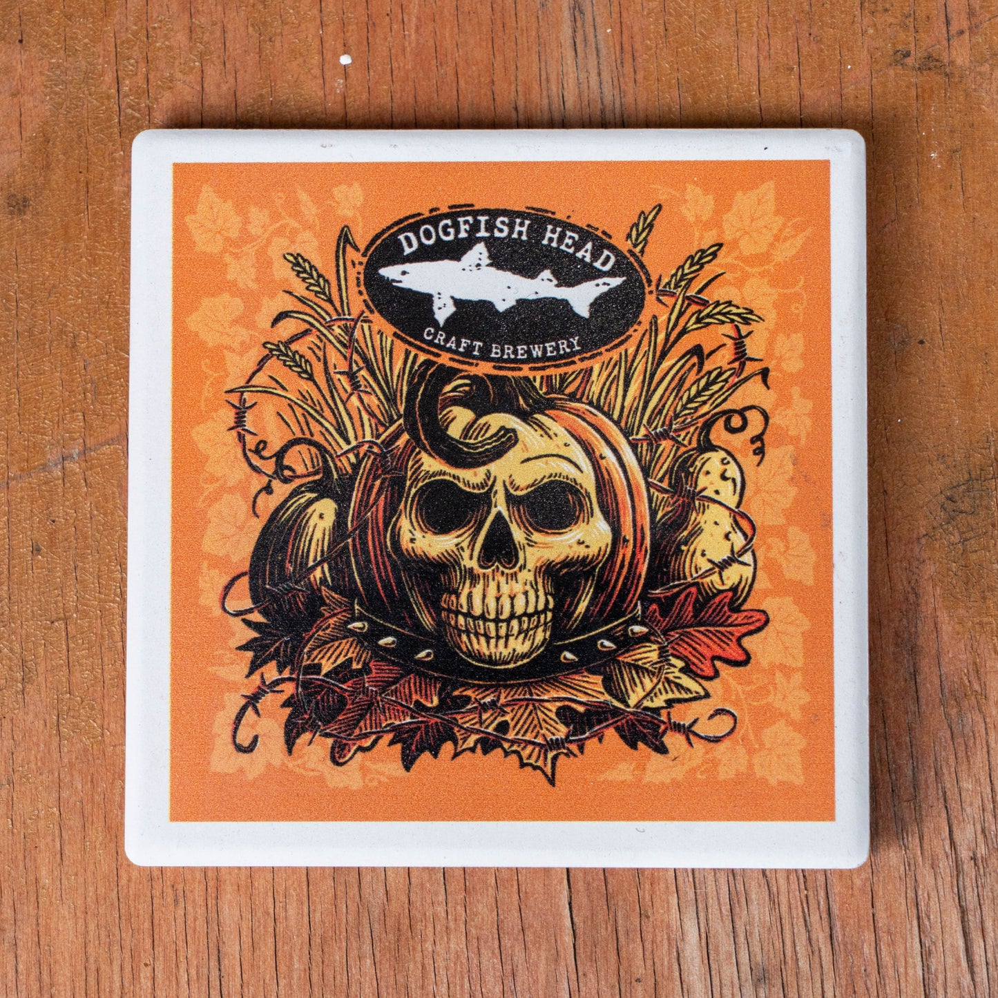 A square coaster with a white border featuring a Halloween-themed illustration of a skull pumpkin, autumn leaves, and the Dogfish Head Craft Brewery logo, placed on a wooden surface.