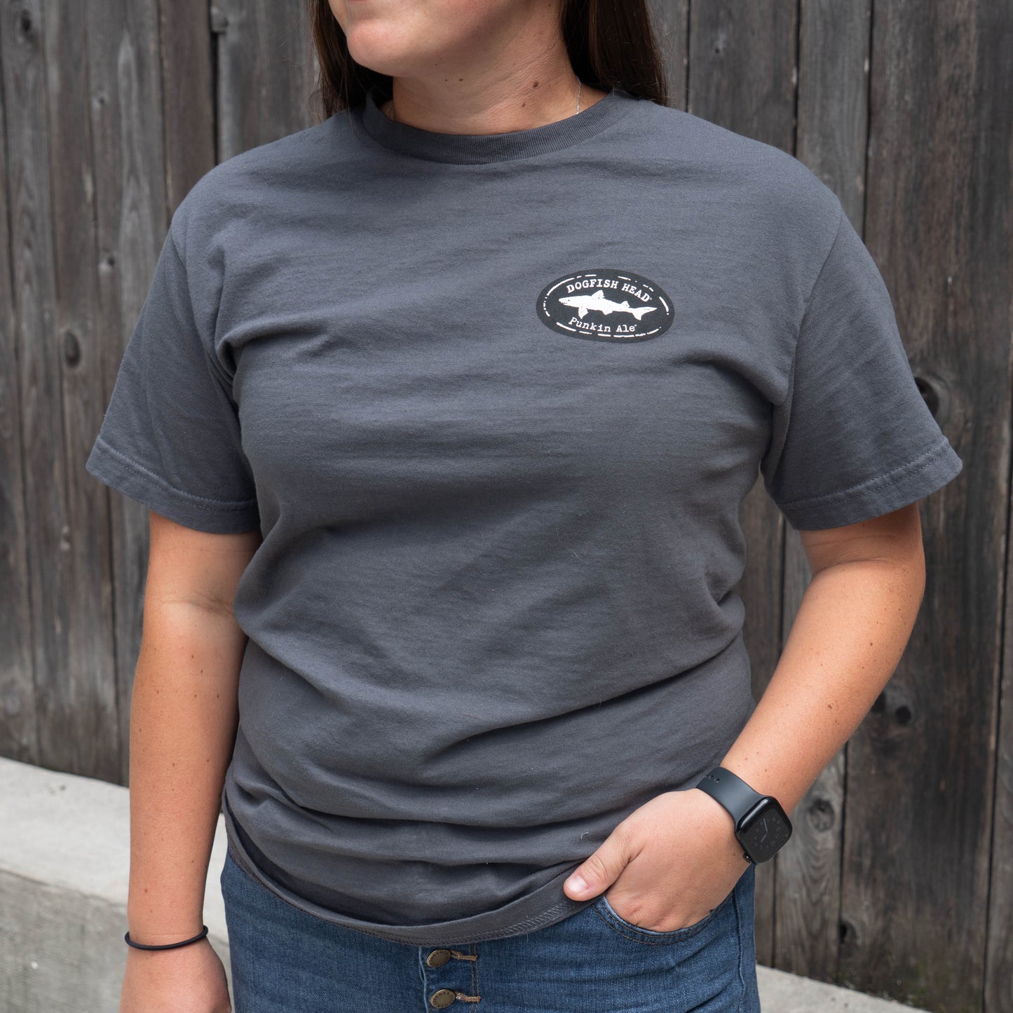 A woman wearing a dark gray crewneck t-shirt with a small black and white circular shark logo on the left chest, standing in front of a weathered wooden fence.