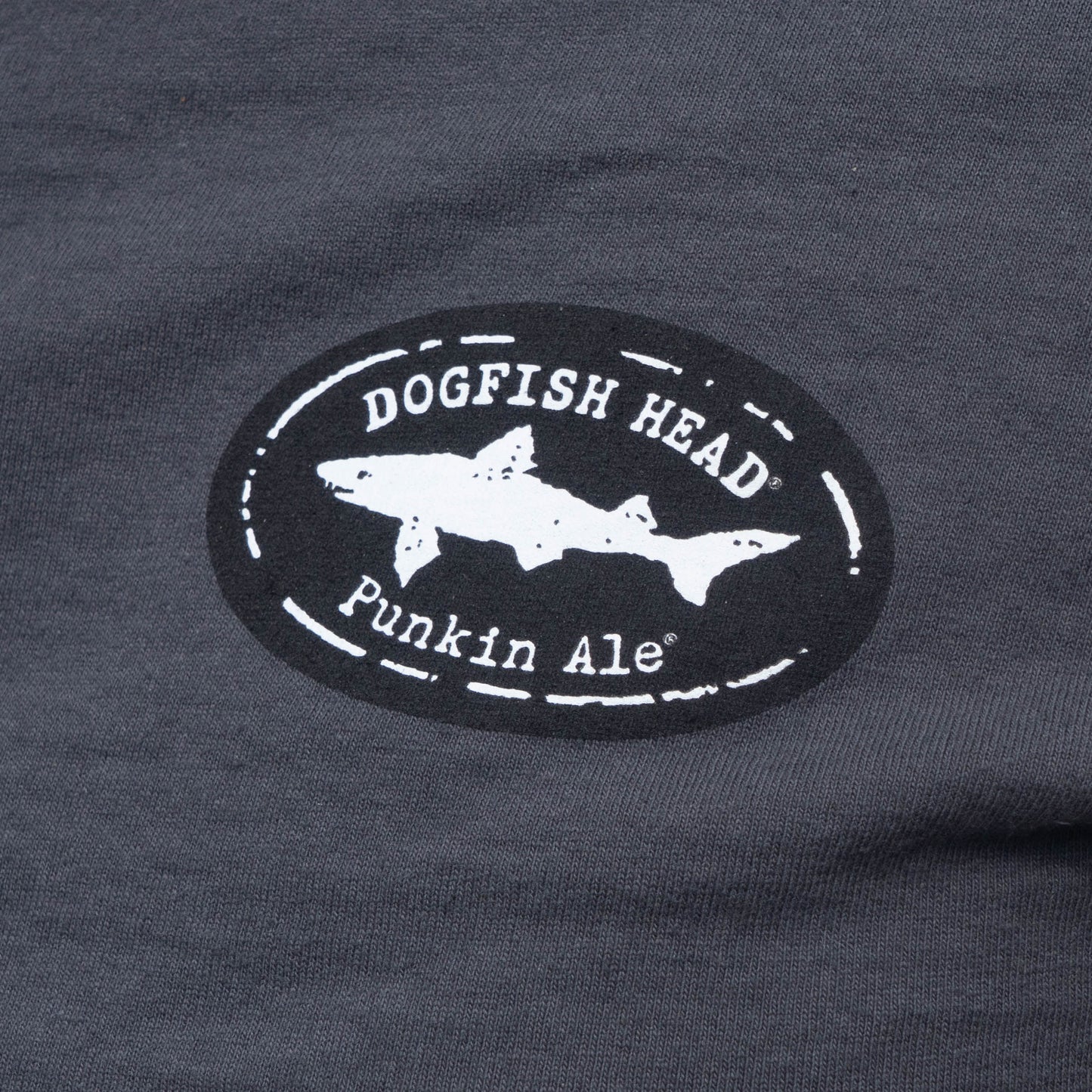 Close-up of a dark charcoal gray t-shirt featuring a circular black and white graphic print on the chest. The graphic displays a white shark silhouette inside a distressed black oval with the text 'DOGFISH HEAD' and 'Punkin Ale'.