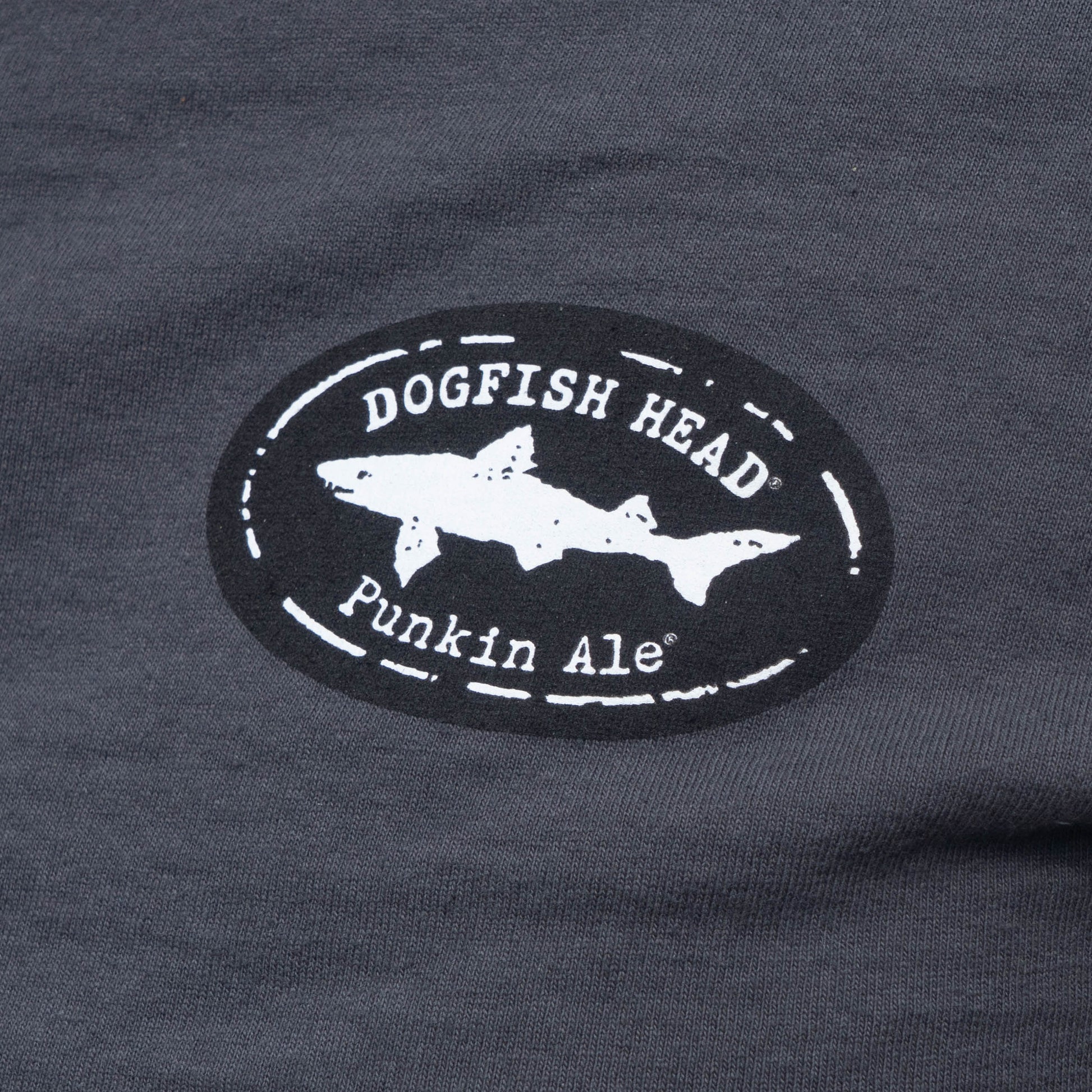 Close-up of a dark charcoal gray t-shirt featuring a circular black and white graphic print on the chest. The graphic displays a white shark silhouette inside a distressed black oval with the text 'DOGFISH HEAD' and 'Punkin Ale'.