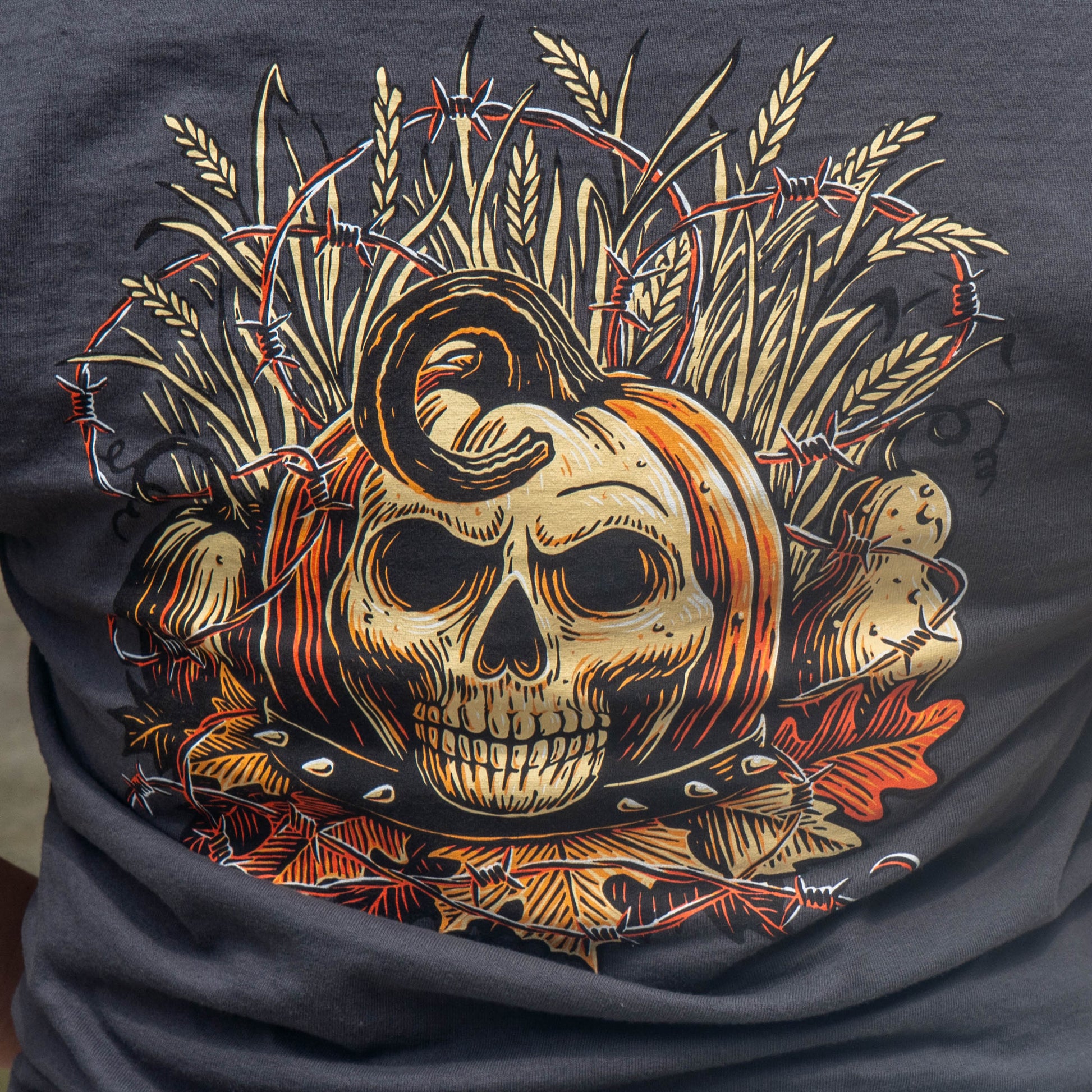 A close-up of a dark charcoal gray t-shirt featuring a detailed graphic print of a skull wrapped in barbed wire, surrounded by wheat stalks, pumpkins, and autumn leaves.