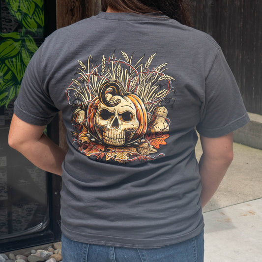 A person viewed from behind wearing a dark gray t-shirt with a large graphic print on the back featuring a skull in a pumpkin, wheat stalks, and autumn leaves. The person is also wearing blue jeans, standing outdoors next to a wooden fence.