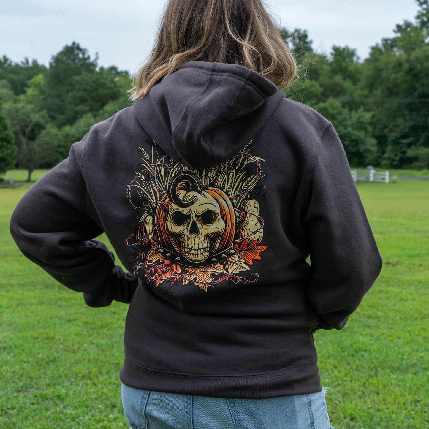 A person standing outdoors in a grassy field, photographed from behind wearing a dark charcoal hoodie with a large graphic print on the back featuring a skull, pumpkin, and autumn leaves.