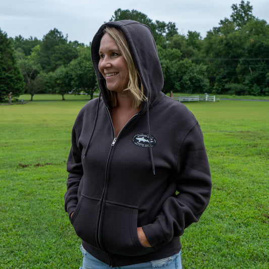 A woman stands smiling with her hands in the pockets of the Punkin Zip Hoodie made from a cotton-recycled polyester blend. The fleece-lined hood adds extra coziness as she stands on green grass, with trees and a white fence visible in the background beneath a cloudy sky.