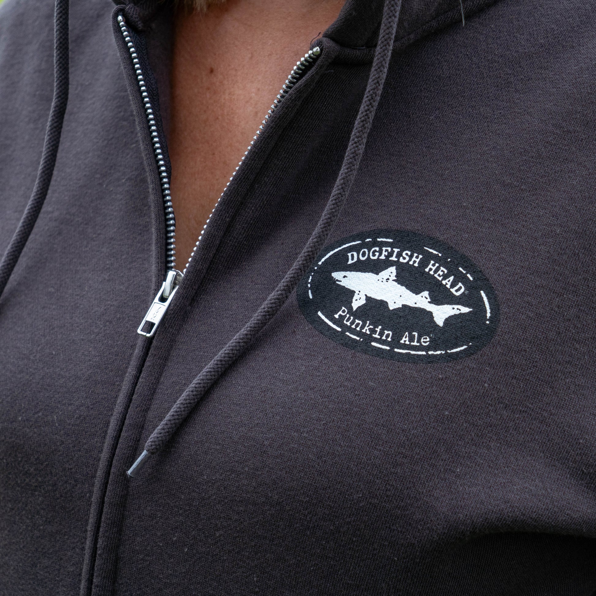 A close-up shows a person wearing the Punkin Zip Hoodie, featuring a Dogfish Head Punkin Ale logo patch on the left chest. The zipper is partially open, with hoodie strings visible. Made from a cotton and recycled polyester blend, this hoodie offers extra comfort with its fleece-lined hood.