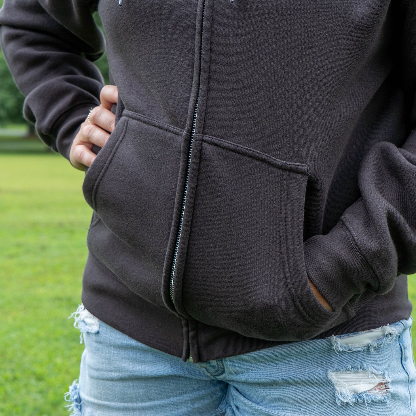 A close-up of a person wearing a dark charcoal zip-up hoodie with front pockets, paired with light blue ripped jeans, standing outdoors on a grassy lawn.