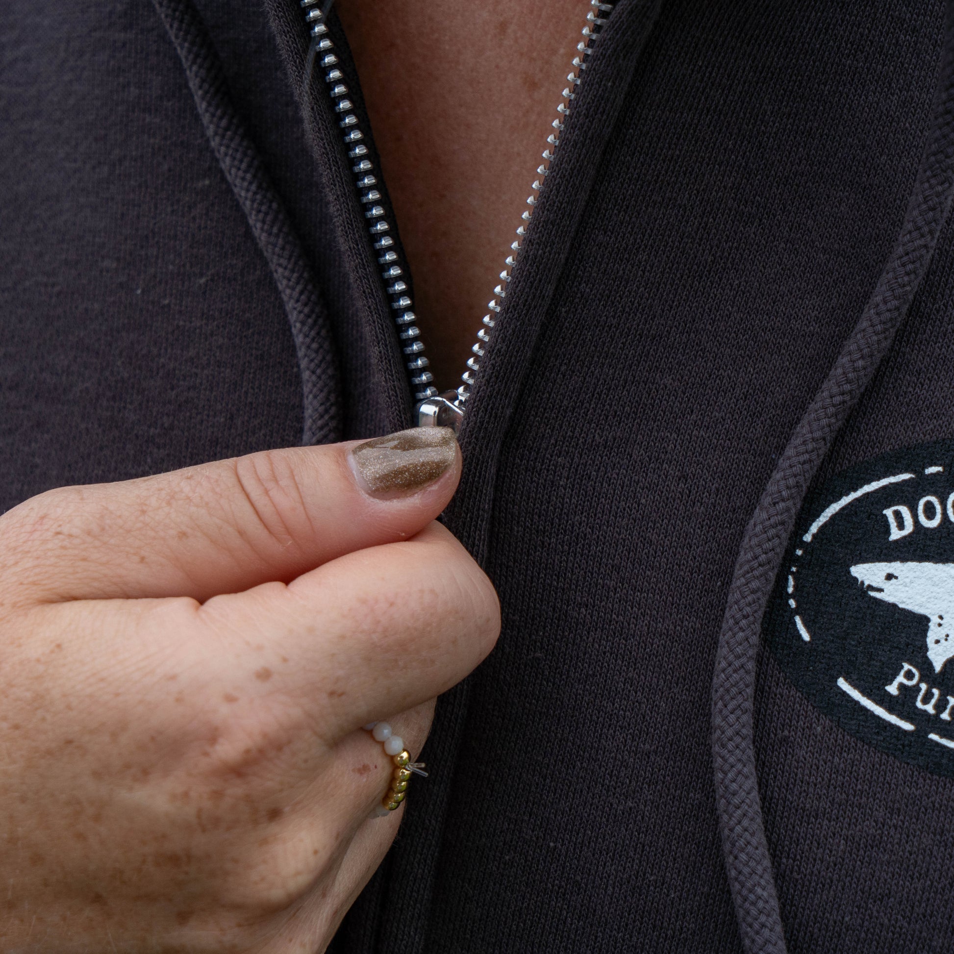 A hand with painted nails and rings is pulling up the zipper on the Punkin Zip Hoodie. Crafted from recycled polyester, this dark zip hoodie features a logo patch with a white dog and partially visible text on the chest.