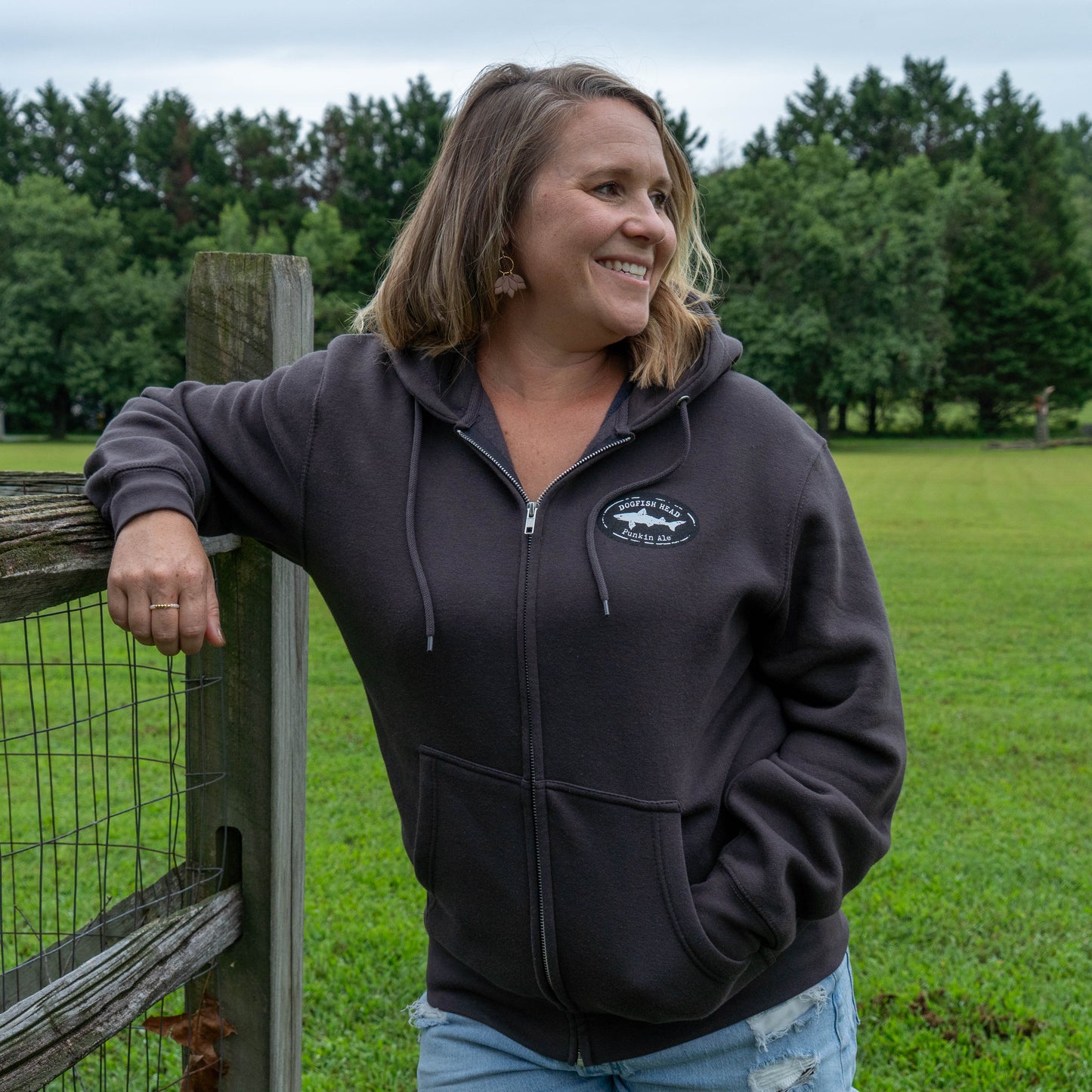 A woman smiling and leaning on a wooden fence in an outdoor grassy field, wearing a dark charcoal zip-up hoodie with a circular Dogfish logo on the left chest.