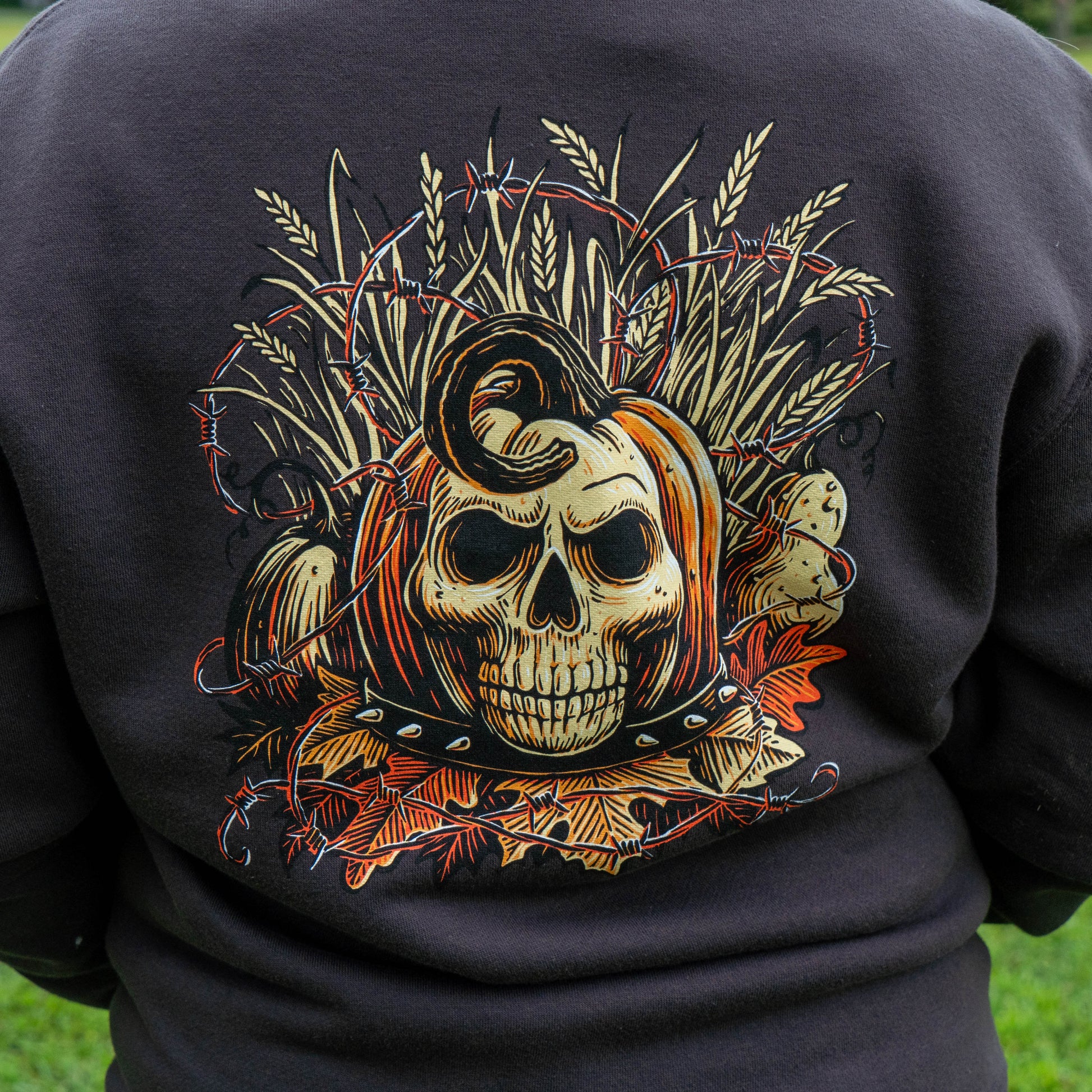 A close-up of the back of a black sweatshirt featuring a large graphic print of a skull surrounded by wheat, barbed wire, and autumn leaves.