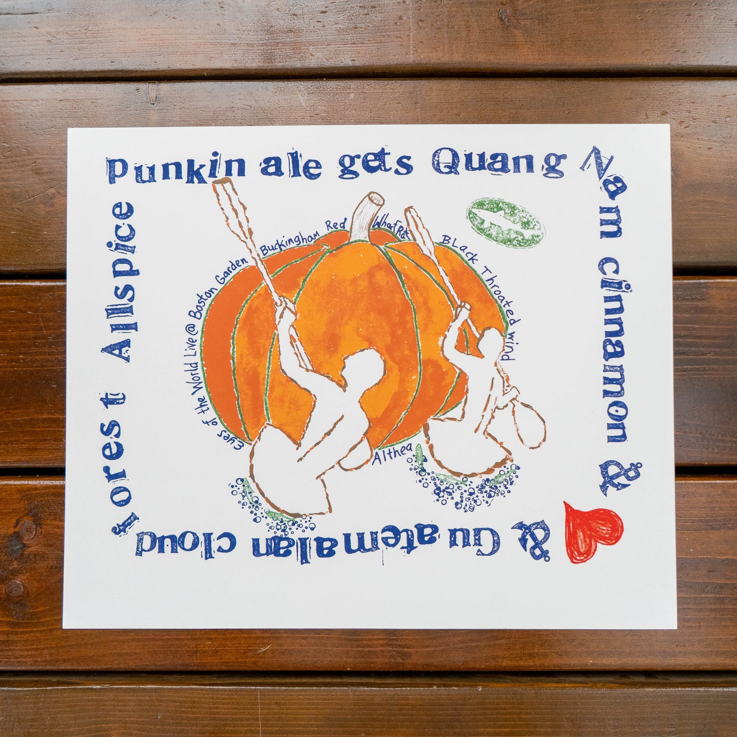 A square white art print featuring a hand-drawn orange pumpkin with two white figures, surrounded by blue and red text, displayed flat on a dark brown wooden surface.