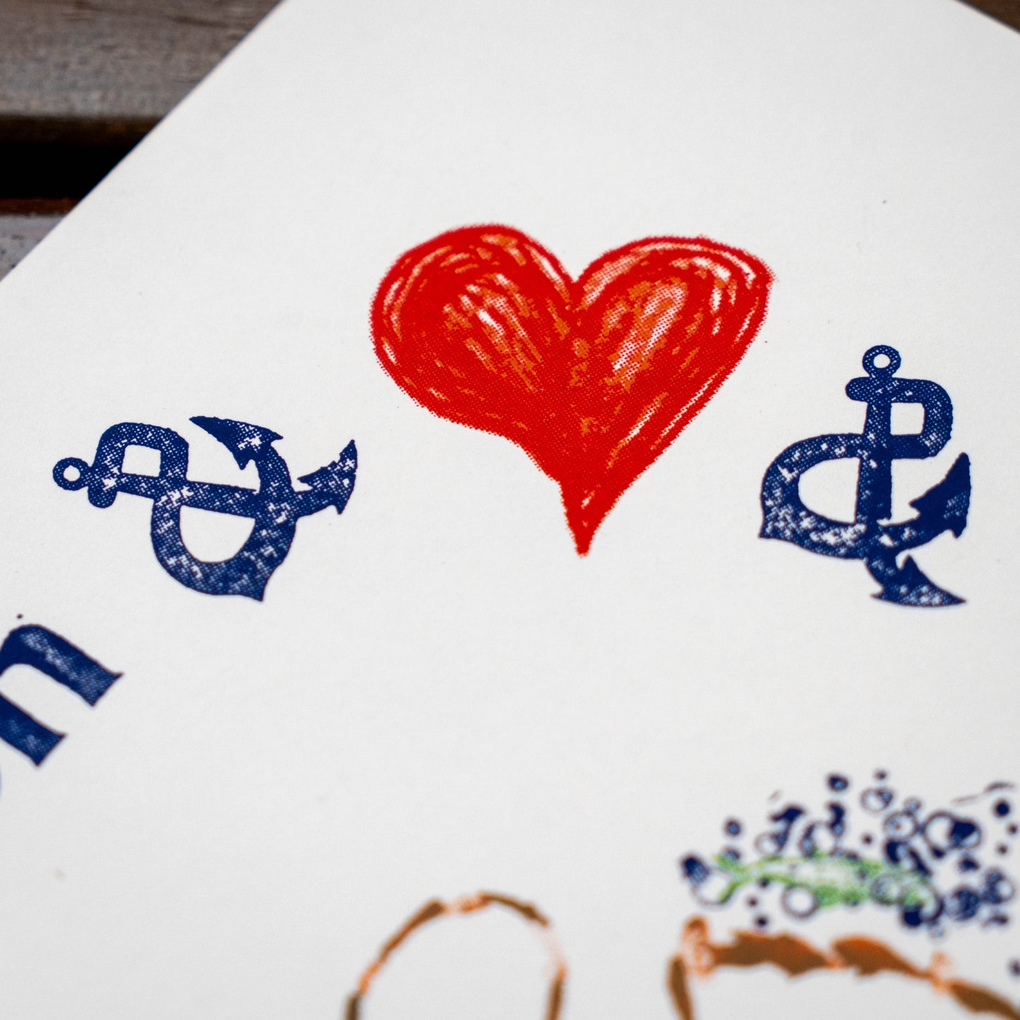 A close-up of a white surface with printed or stamped anchor motifs in dark blue and a red heart in the center. Part of another blue graphic and a small multicolor mark are visible at the bottom edges.