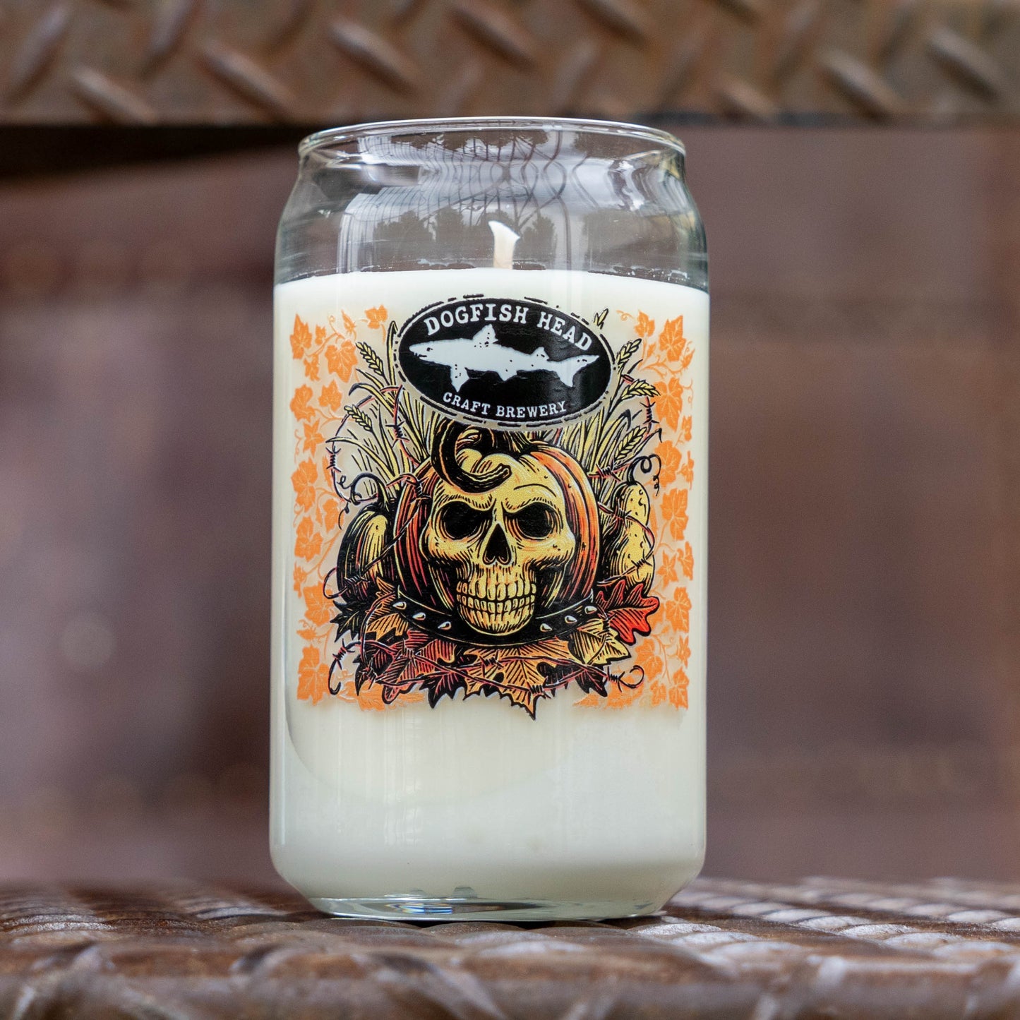 A clear glass jar candle with white wax and a printed front label reading Dogfish Head Craft Brewery. The label features a skull, pumpkin, and orange autumn leaf design.
