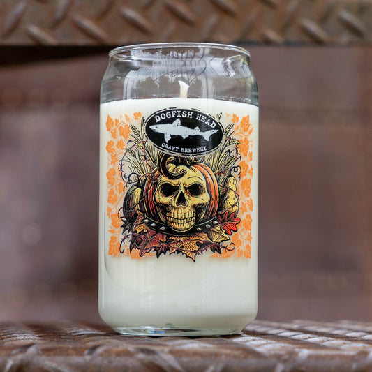 A clear glass jar candle with white wax and a printed front label reading Dogfish Head Craft Brewery. The label features a skull, pumpkin, and orange autumn leaf design.