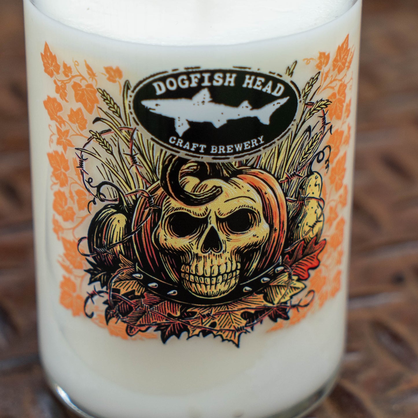 A close-up of a Dogfish Head Craft Brewery glass with a Halloween-themed label featuring a skull, pumpkins, autumn leaves, and a shark logo, filled with an off-white beverage.