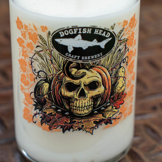 A close-up of a Dogfish Head Craft Brewery glass with a Halloween-themed label featuring a skull, pumpkins, autumn leaves, and a shark logo, filled with an off-white beverage.