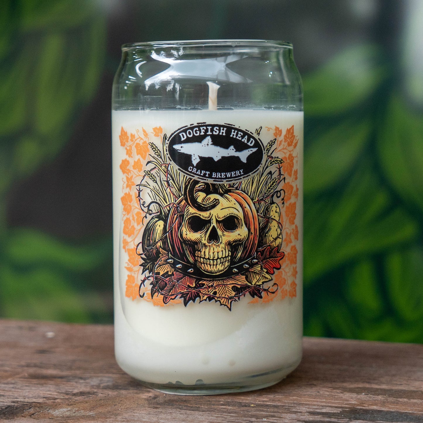 A clear glass jar candle with white wax sits on a wooden surface. The front label reads 'DOGFLISH HEAD CRAFT BREWERY' and features a skull over a pumpkin with orange autumn leaves.