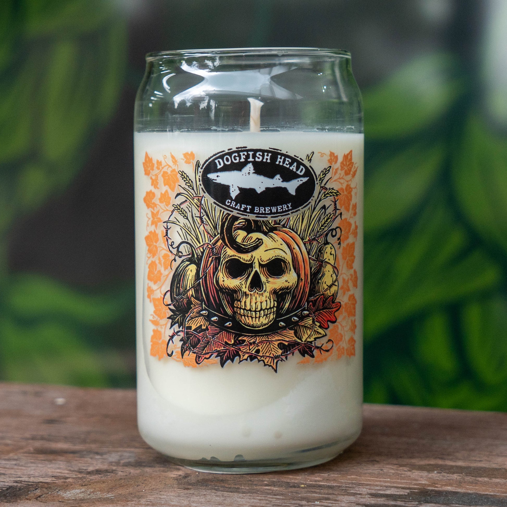 A clear glass jar candle with white wax sits on a wooden surface. The front label reads 'DOGFLISH HEAD CRAFT BREWERY' and features a skull over a pumpkin with orange autumn leaves.