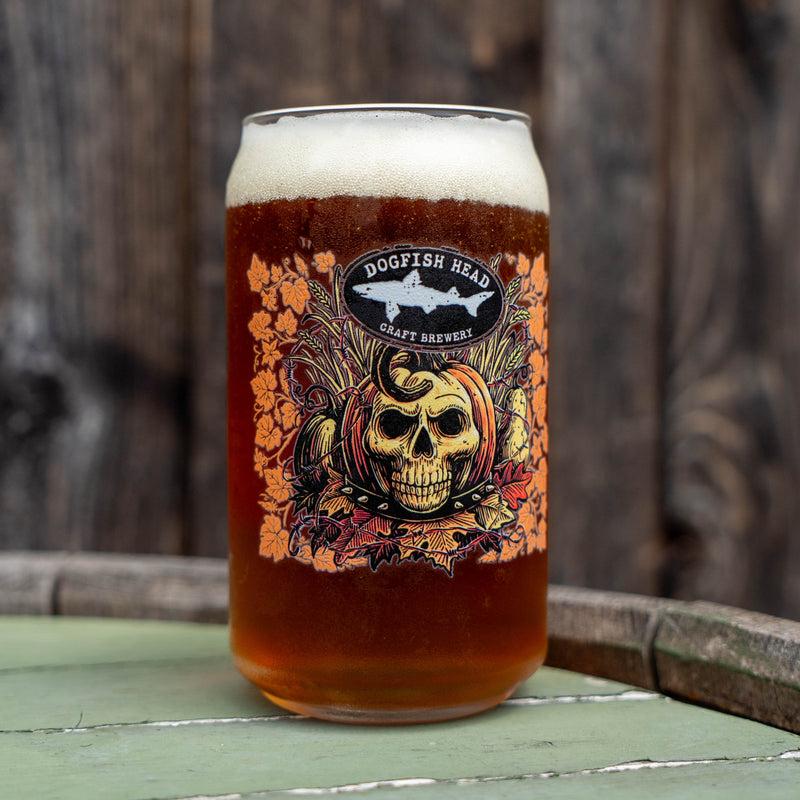 A glass of Dogfish Head Craft Brewery IPA beer with a thick head of white foam, featuring a decorative label with a skull, shark, and autumn leaves, set on a rustic wooden table.