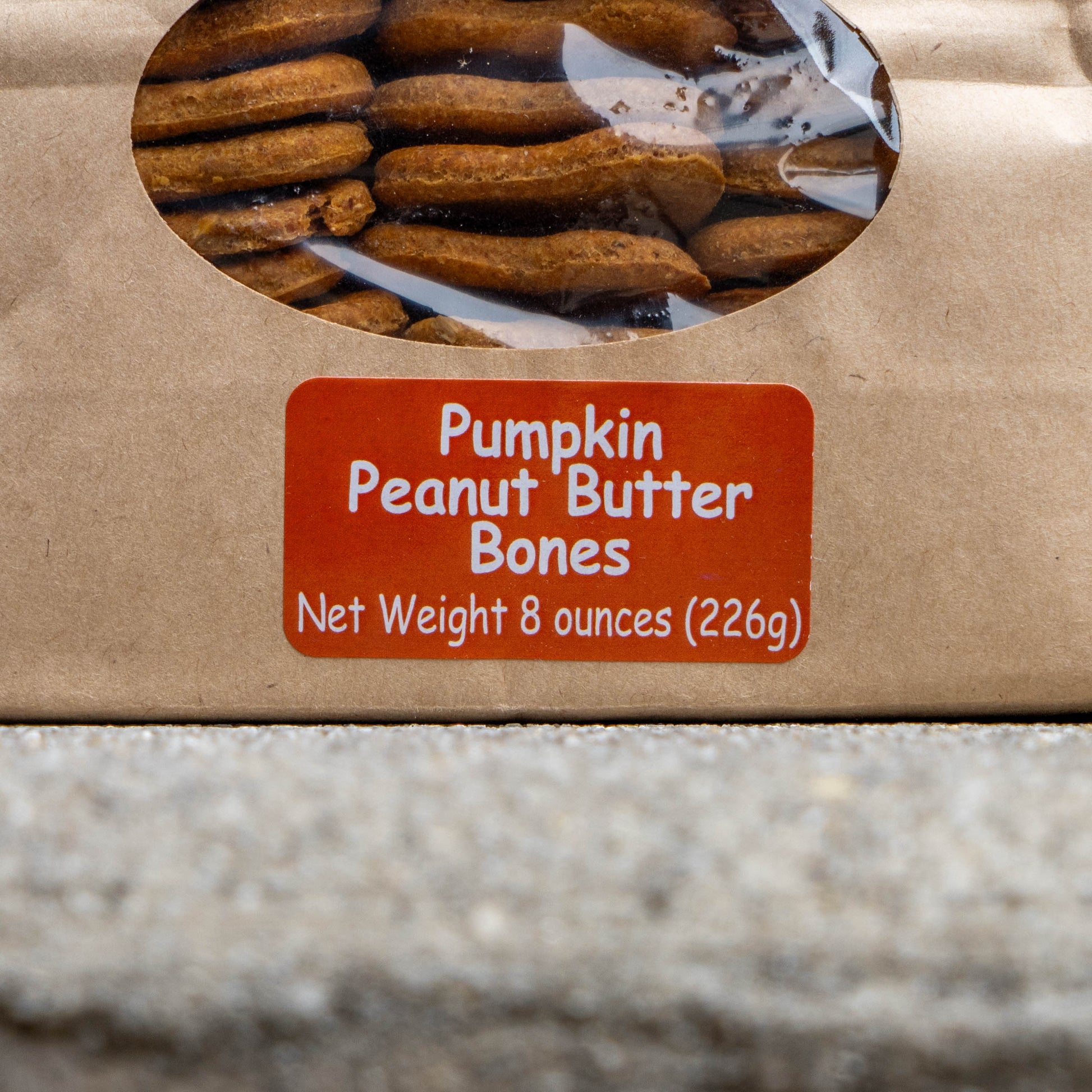 A close-up of a brown kraft paper bag containing Pumpkin Peanut Butter Bones dog treats, with a clear oval window showing the treats inside and a red label displaying the product name and net weight.