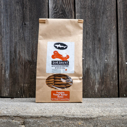 A bag of Dog Bark! Pumpkin Peanut Butter Bites dog treats standing upright on a concrete surface against a dark weathered wood plank background.