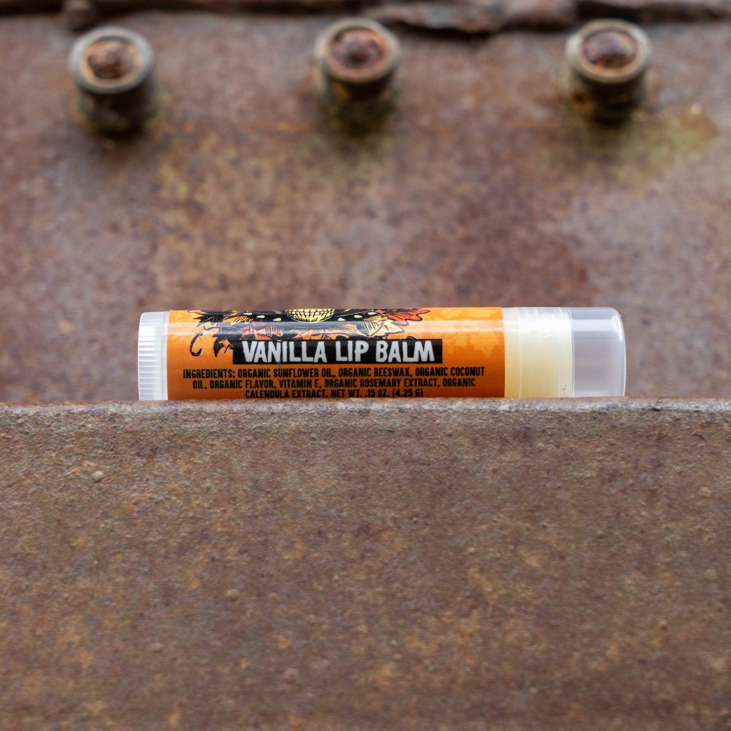 A tube of Vanilla Lip Balm by The Lip Balm Company resting horizontally on a textured, rusty metal surface with three rusty bolts in the background.