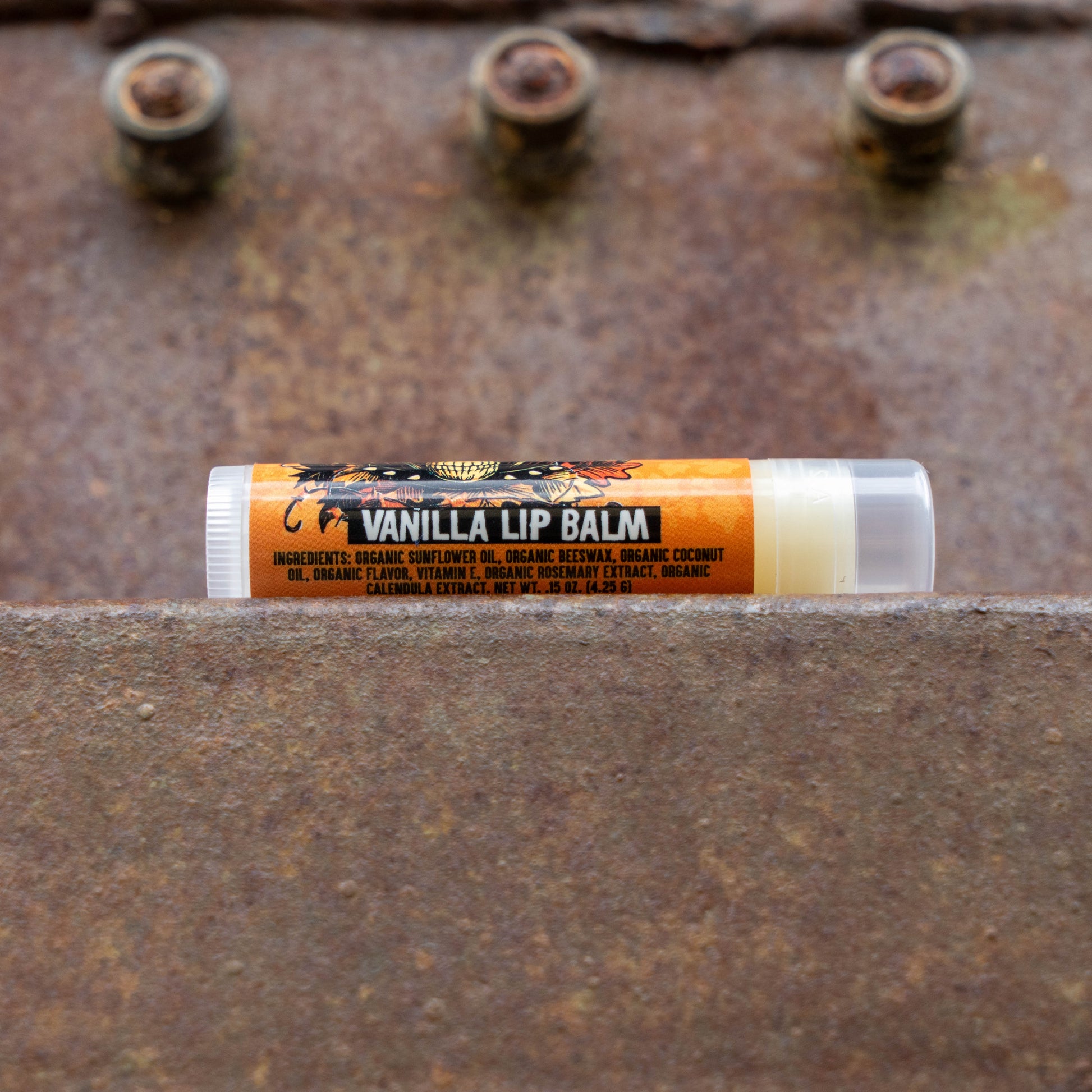 A tube of Vanilla Lip Balm by The Lip Balm Company resting horizontally on a textured, rusty metal surface with three rusty bolts in the background.