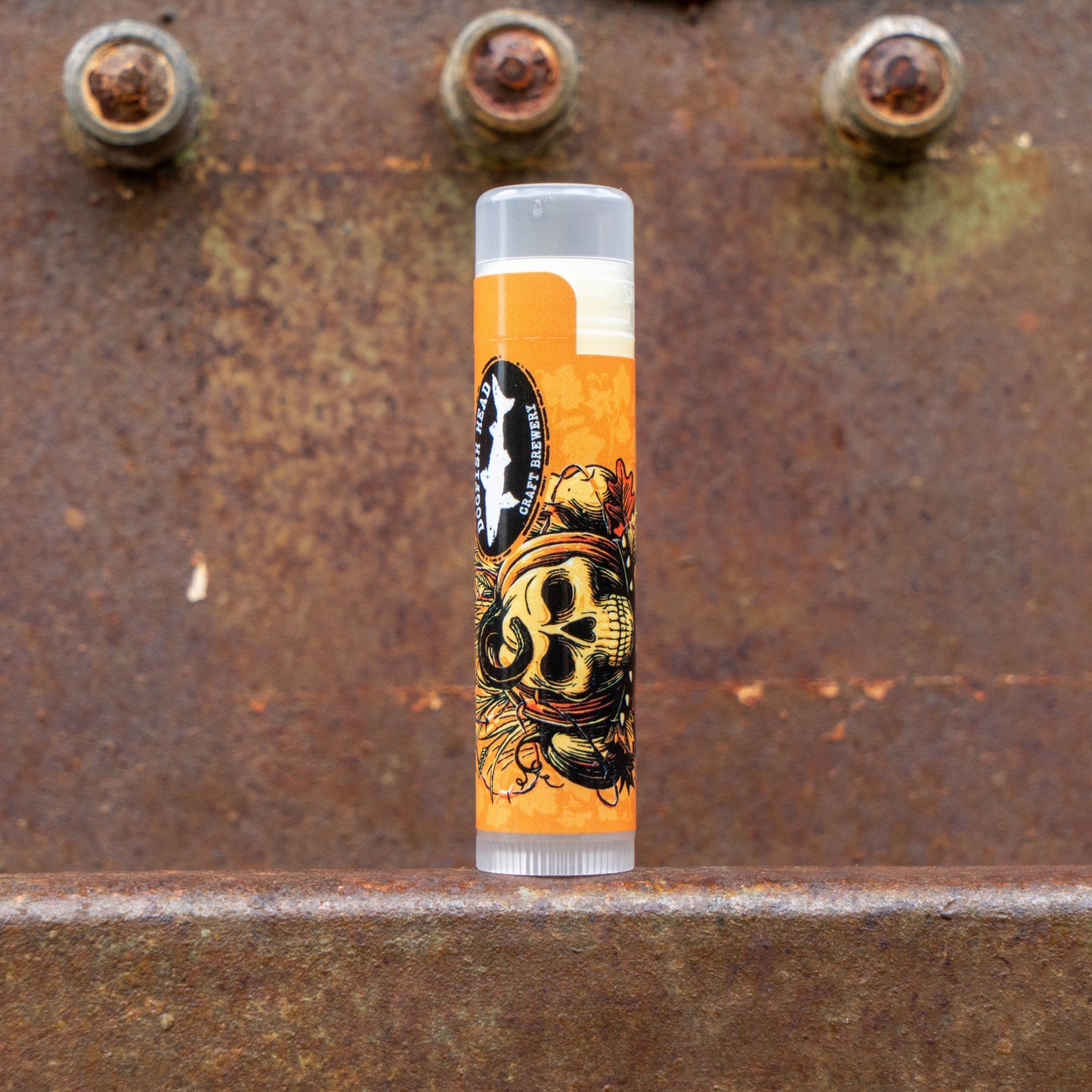 A single lip balm tube standing upright on a rusty metal surface, with a bright orange label featuring a skull illustration and a circular black-and-white logo. The background is a textured rusted metal surface with visible bolts.