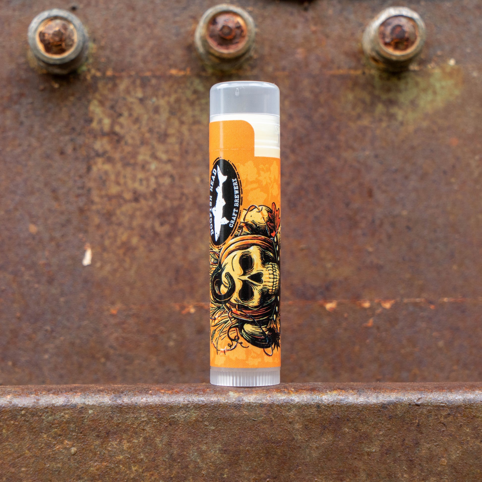 A single lip balm tube standing upright on a rusty metal surface, with a bright orange label featuring a skull illustration and a circular black-and-white logo. The background is a textured rusted metal surface with visible bolts.