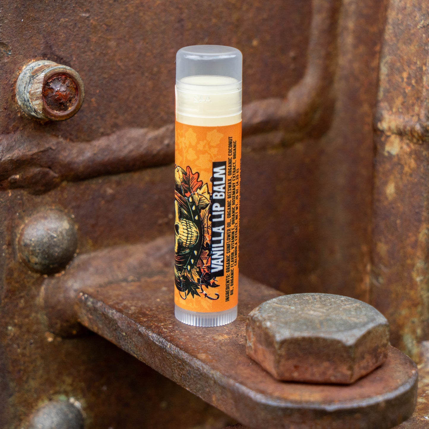 A single tube of lip balm standing upright on a rusty metal surface. The tube has a clear cap, a cream-colored balm visible at the top, and an orange label with black text reading 'VANILLA LIP BALM' and a decorative illustration.