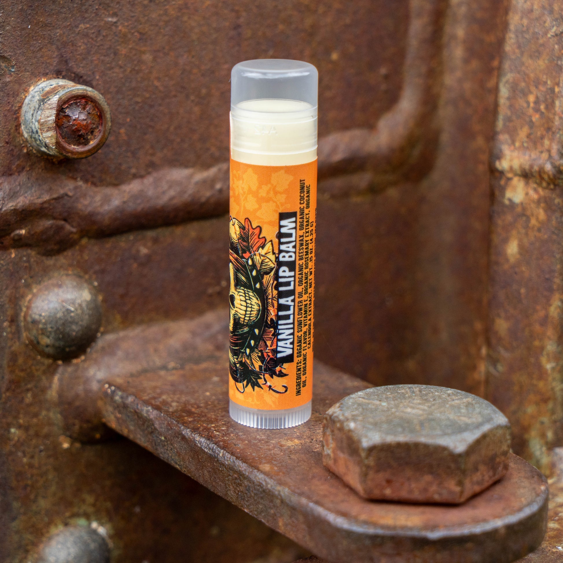 A single tube of lip balm standing upright on a rusty metal surface. The tube has a clear cap, a cream-colored balm visible at the top, and an orange label with black text reading 'VANILLA LIP BALM' and a decorative illustration.