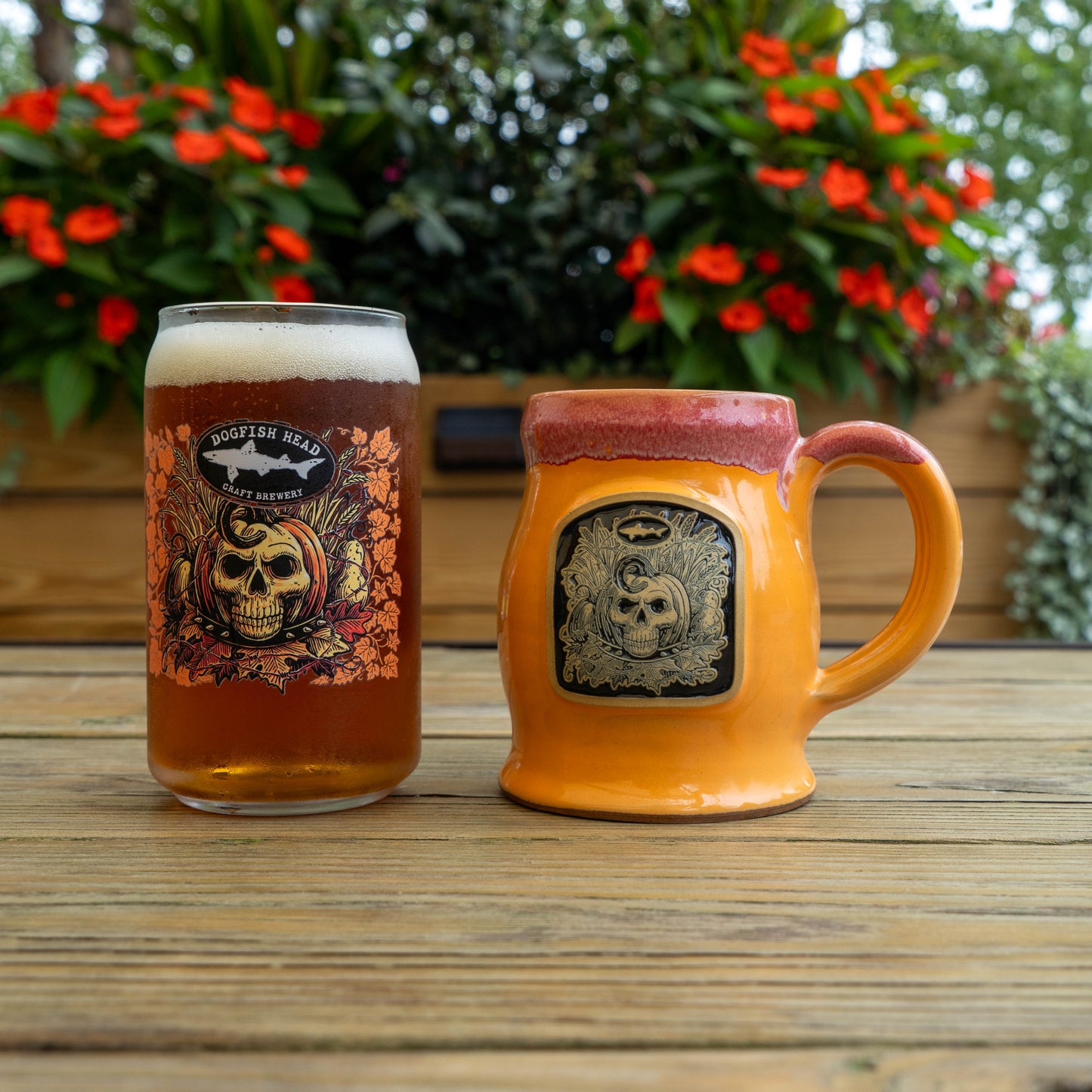 A clear glass mug filled with amber beer and a ceramic orange beer stein with a skull graphic, placed on a wooden table with flowers and greenery in the background.