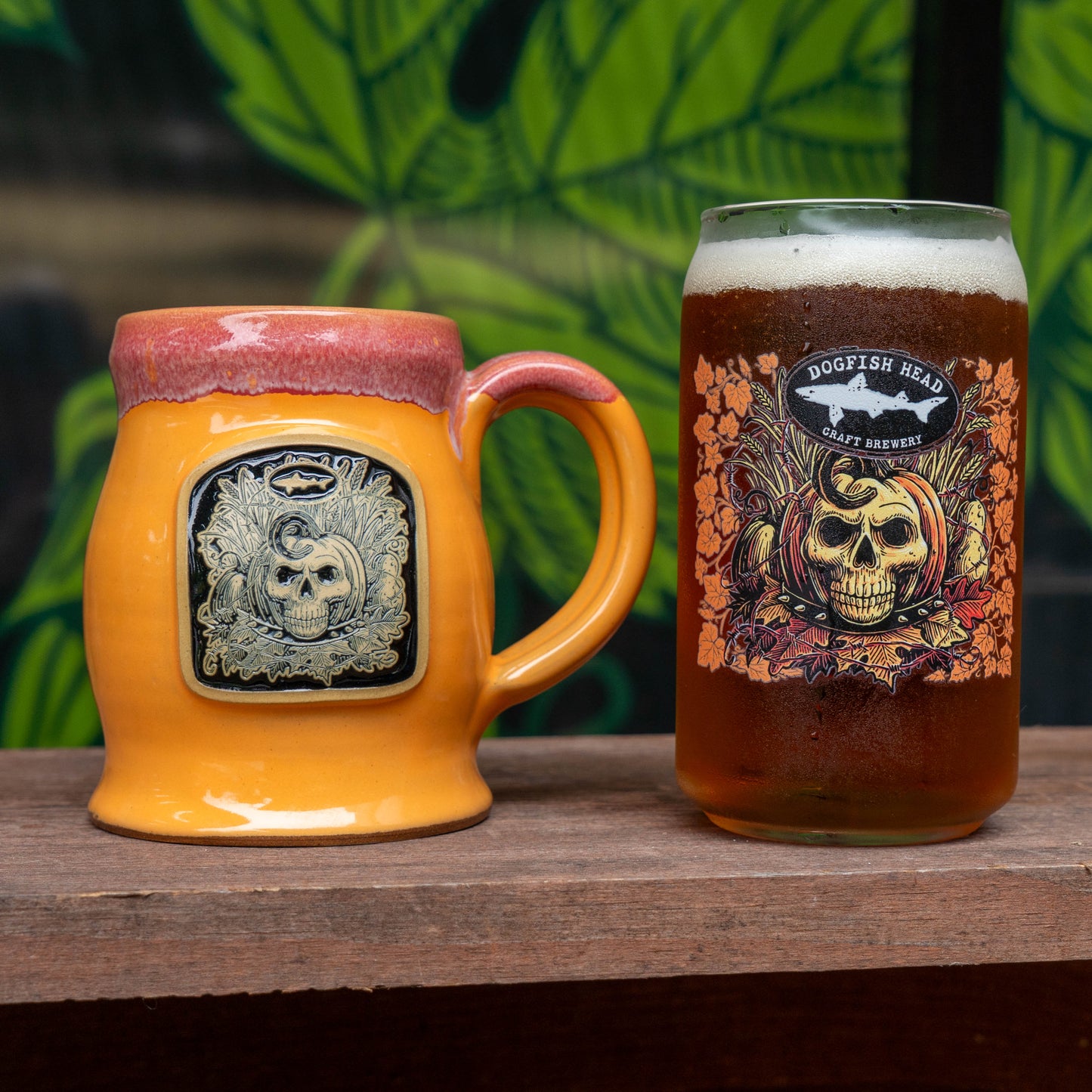 A ceramic orange mug with a skull and pumpkin design and a dripping pinkish-brown glaze, shown next to a glass of amber beer with a 'Dogfish Head Craft Brewery' label, set on a wooden surface with a green leafy background.