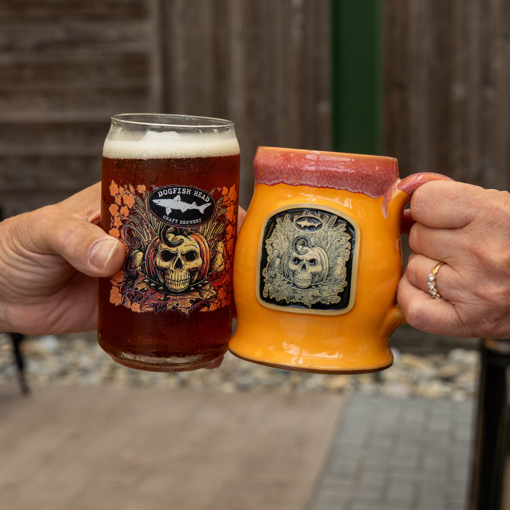 Two hands holding a clinking pair of beer glasses outdoors. The left glass is a clear pint glass with a Dogfish Head Craft Brewery Halloween-themed label, filled with amber beer and topped with white foam.