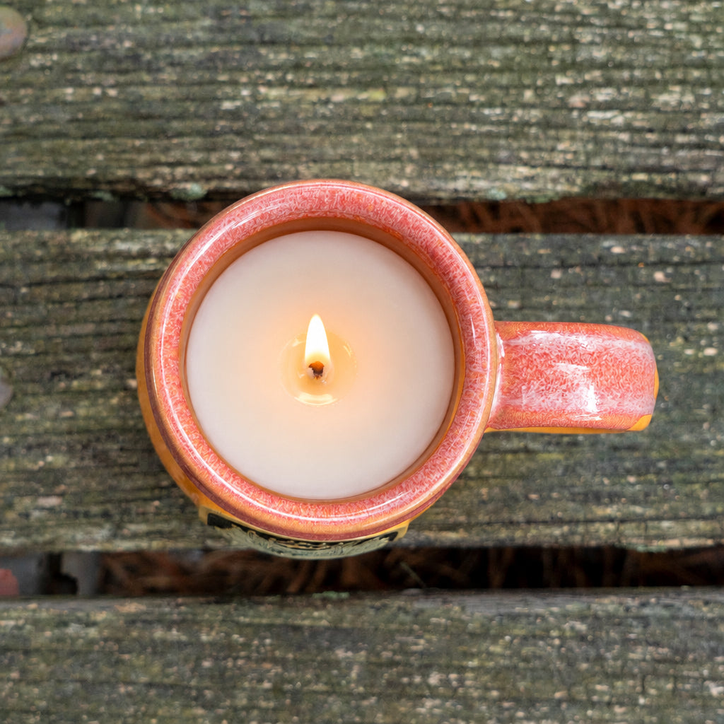 Ceramic Mug candle with white wax interior with orange and red glaze on wooden table.