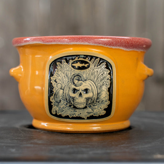 A ceramic cauldron-shaped planter in bright orange with a reddish-pink glazed rim, featuring a black decorative panel on the front with a skull and autumn leaf design.