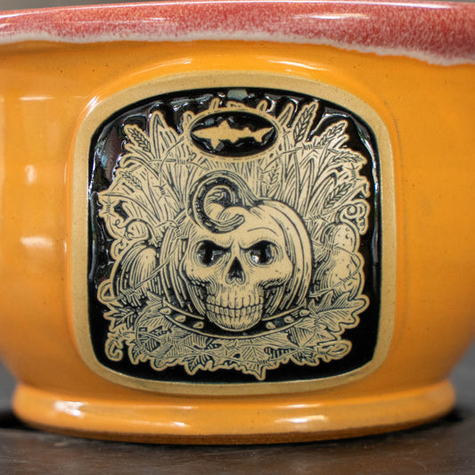 Close-up of a glossy orange ceramic bowl featuring a raised black and beige Halloween-themed relief design on the front with a skull, pumpkins, and autumn leaves.