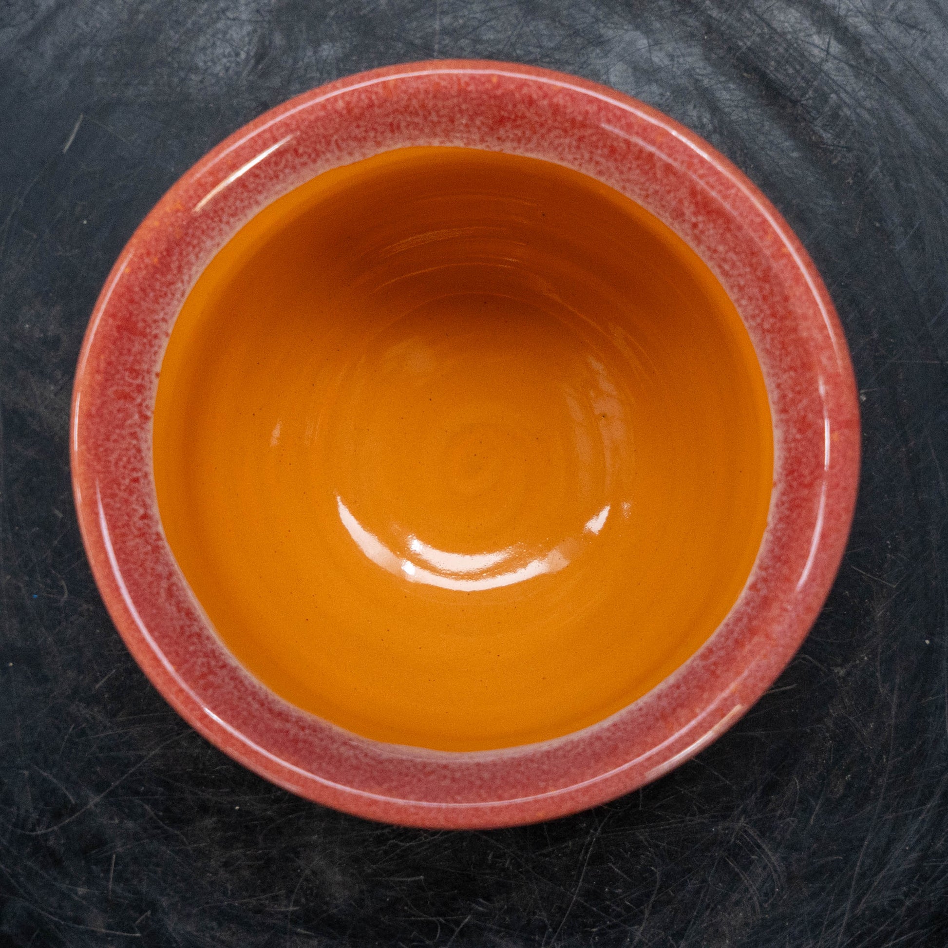 A ceramic bowl with a glossy orange interior and a speckled terracotta red exterior, shown from a top-down view on a dark textured background.
