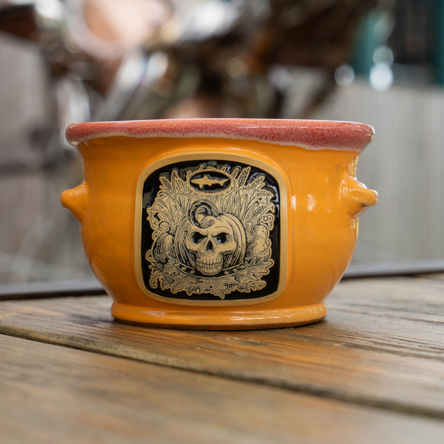 An orange ceramic crock with a Halloween-themed black and gold skull plaque on the front, resting on a wooden surface with a blurred background.