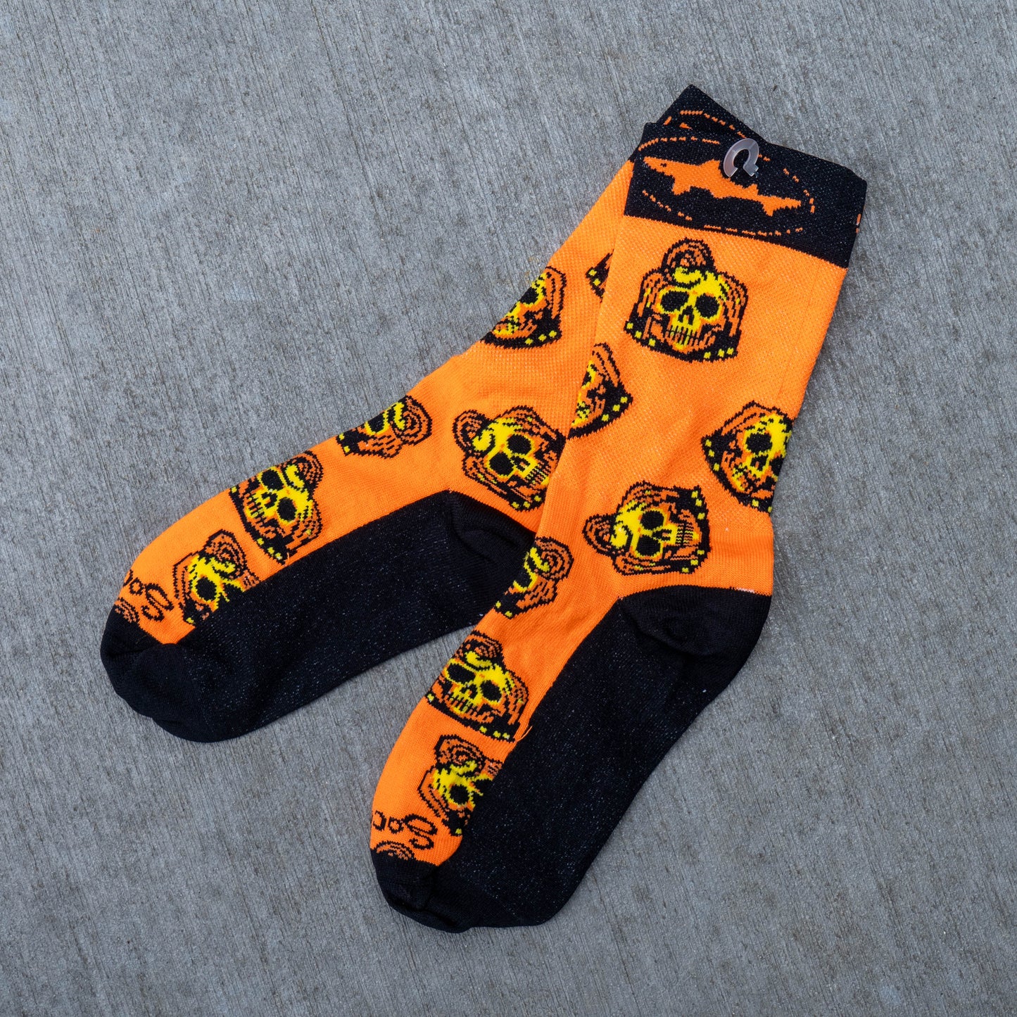 A pair of orange and black crew socks with a repeating pumpkin skull pattern, laid flat on a gray textured fabric background.