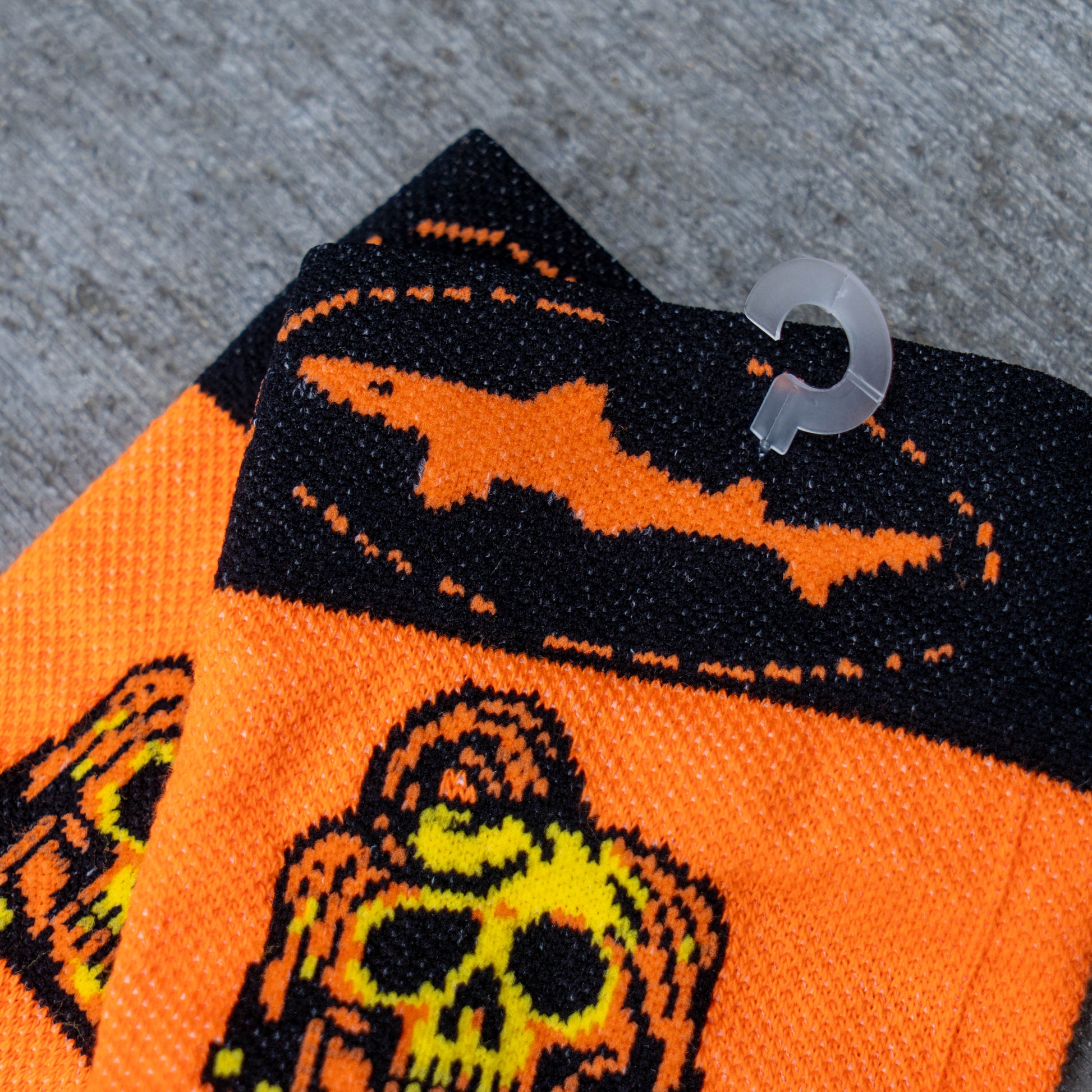 Bright orange knit socks with black and yellow Halloween-themed graphics, including a skull and a bat silhouette, shown folded on a gray textured surface with a clear plastic hanger hook visible.