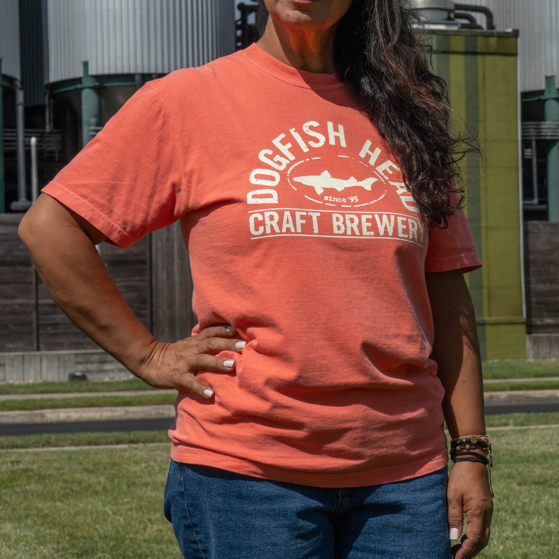 A woman wearing a coral orange graphic t-shirt that reads 'DOGFISH HEAD CRAFT BREWERY' with a shark logo, standing outdoors on a grassy lawn with industrial buildings in the background.