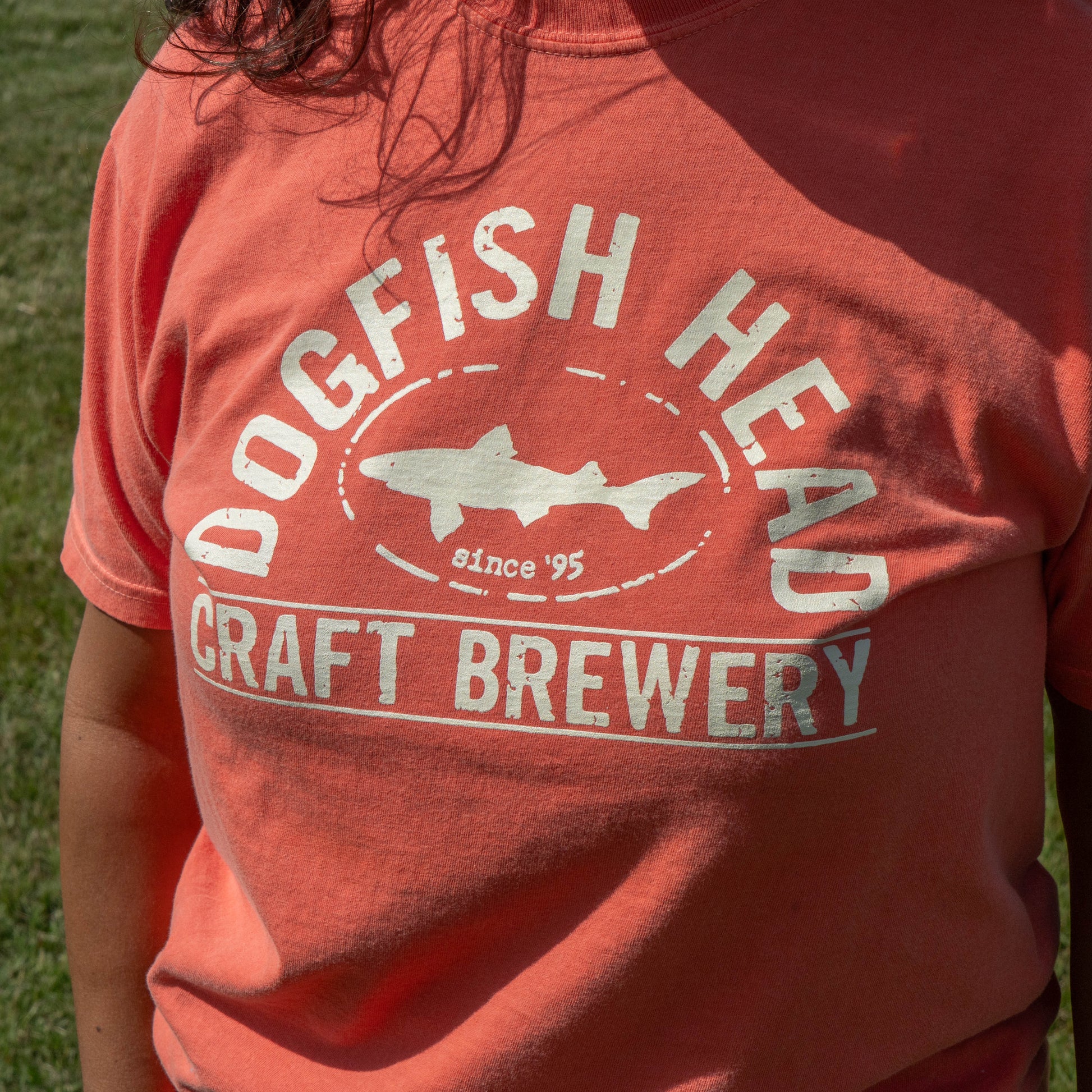 A close-up of a person wearing a coral orange t-shirt with a distressed white graphic that reads 'DOGFISH HEAD CRAFT BREWERY since '95' with a fish silhouette.