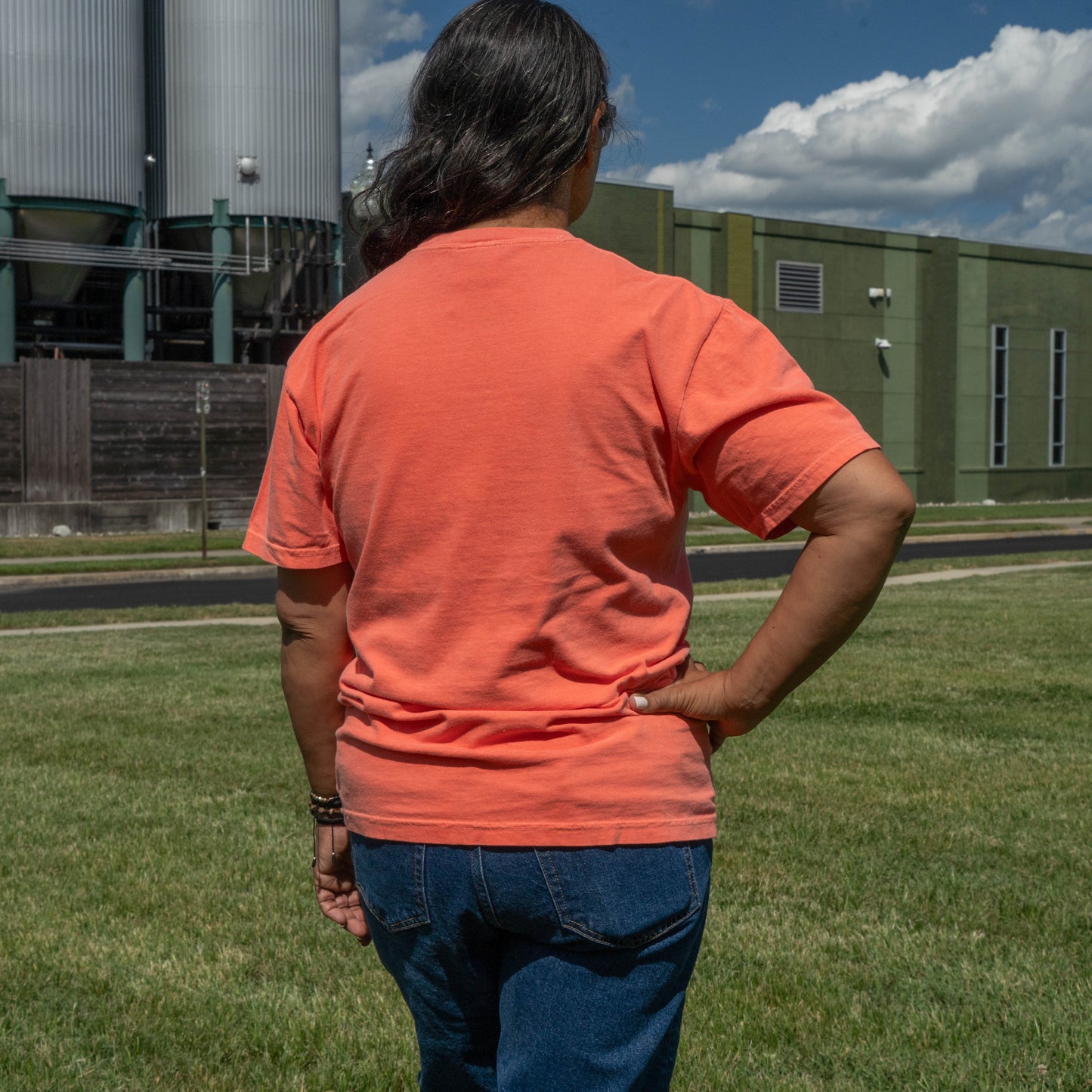 A person viewed from behind wearing a short-sleeve crewneck t-shirt in a bright coral-orange color, paired with blue jeans, standing on a grassy field with an industrial building and sky in the background.
