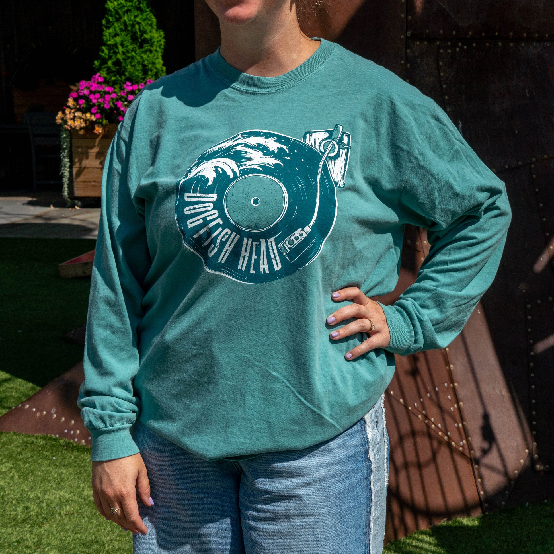 A person wearing a teal long-sleeve graphic T-shirt with a vinyl record design that reads 'Dogfish Head' and light blue jeans, standing outdoors in front of a rustic brown wall and green grass.