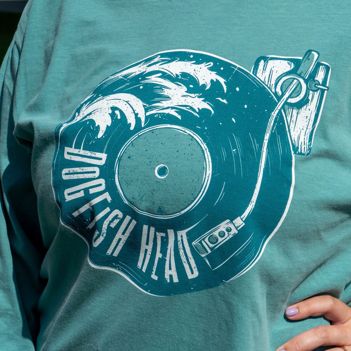 A close-up of a person wearing a teal long-sleeve t-shirt with a large white graphic print of a vinyl record and the words 'DOGFISH HEAD'.