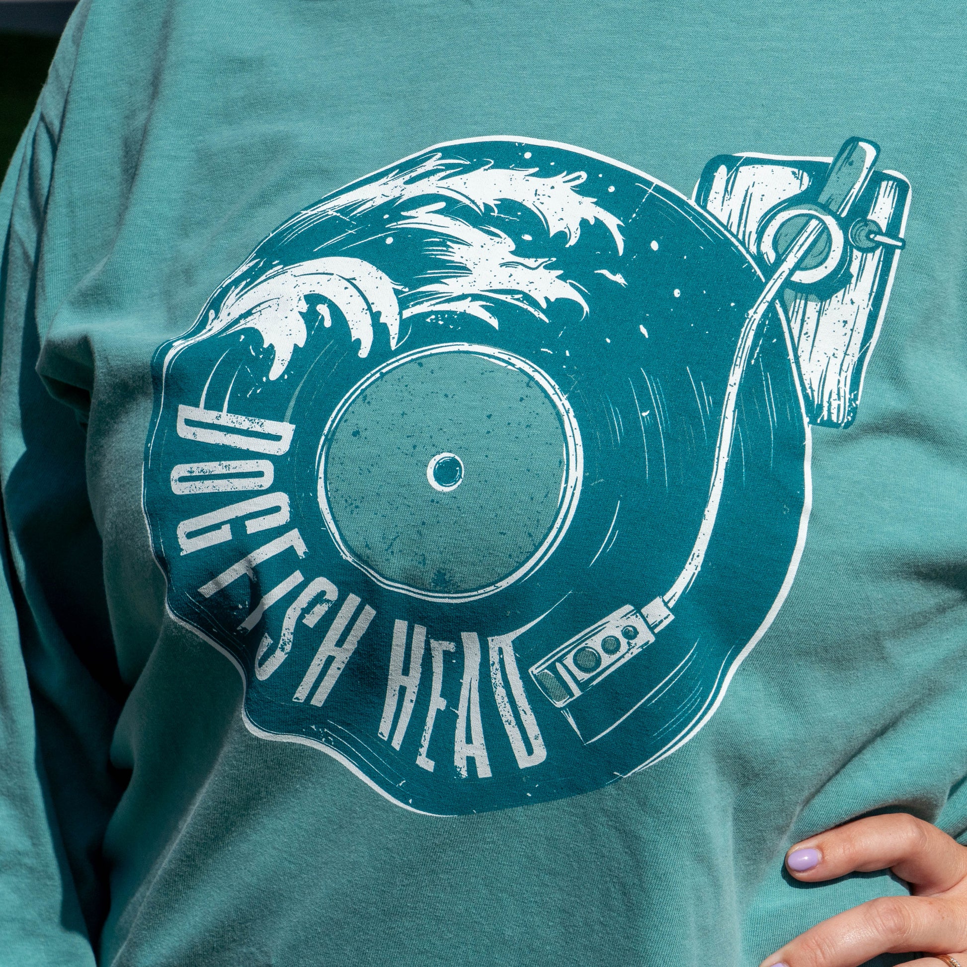 A close-up of a person wearing a teal long-sleeve t-shirt with a large white graphic print of a vinyl record and the words 'DOGFISH HEAD'.