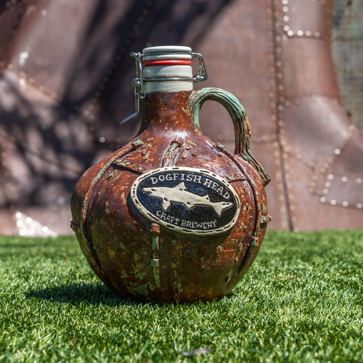 A rustic, weathered Steampunk Punkin Ceramic Growler by Romanick Pottery with a metal clasp sits on green grass, set against a metallic, riveted background.
