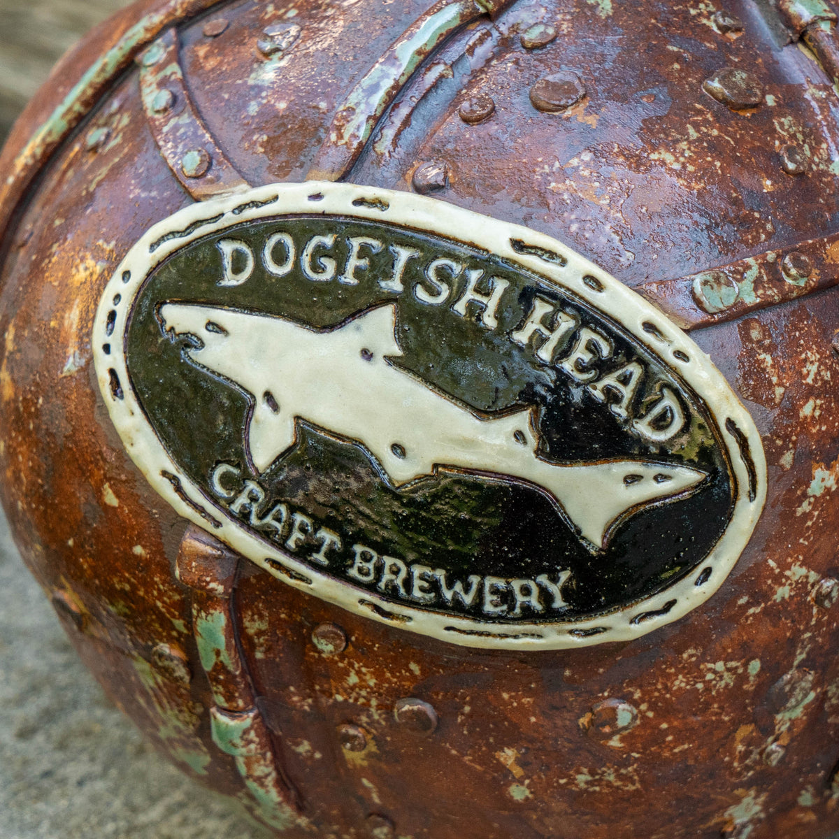 A weathered metal surface features an oval plaque with a shark silhouette and the text Dogfish Head Craft Brewery embossed on it, evoking the artisanal spirit behind Romanick Pottery’s Steampunk Punkin Ceramic Growler.