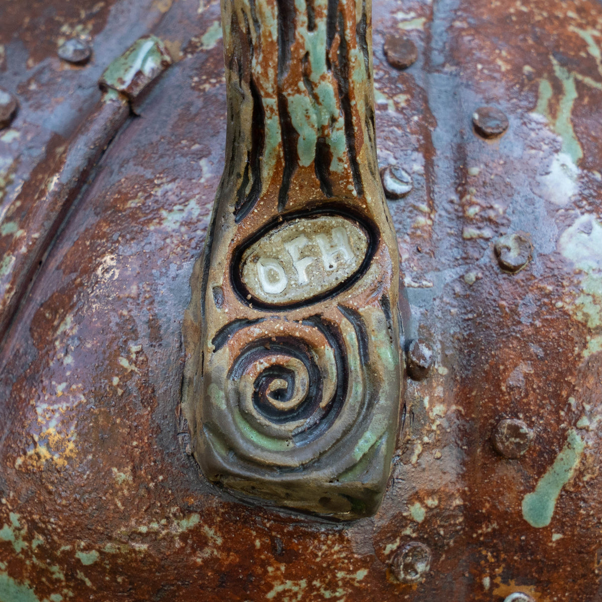 Close-up of the Steampunk Punkin Ceramic Growler by Romanick Pottery, showcasing its textured, weathered metal-like ceramic surface with a distinctive handle. The handle, reminiscent of those found on limited edition growlers, features a spiral pattern and is engraved with the letters “OFH.” The surface displays touches of rust, green patina, and several decorative rivets.
