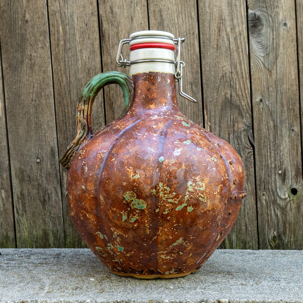 A rustic, pumpkin-shaped Steampunk Punkin Ceramic Growler from Romanick Pottery, featuring a green handle and a metal clasp top, sits on a concrete ledge in front of a weathered wooden fence. This unique piece is one of their handmade ceramic growlers, perfect for collectors and enthusiasts alike.